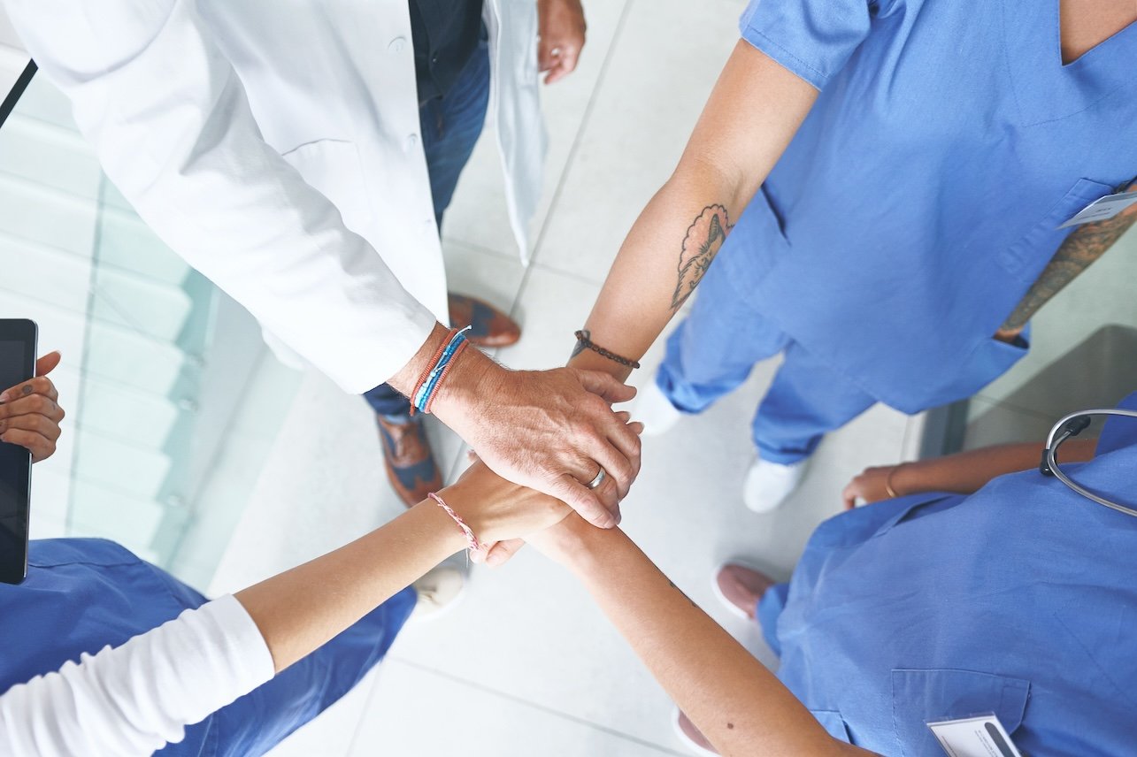 Hands together, above and meeting with doctors in hospital for solidarity, synergy and healthcare. Medical, teamwork and commitment with people in clinic for insurance, community and collaboration